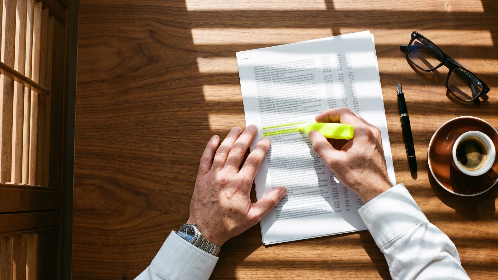 Vista dall'alto di due mani che evidenziano in lime alcune righe di un documento stampato su una scrivania di noce, accanto a occhiali, penna stilografica e caffè: la rilettura della minuta del mutuo prima del rogito